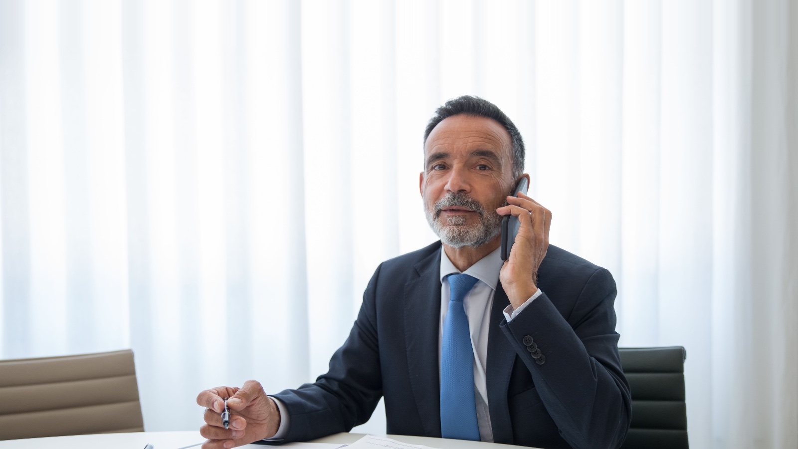 Company director in a suit taking a phone call at a boardroom table with policy paperwork in front of him